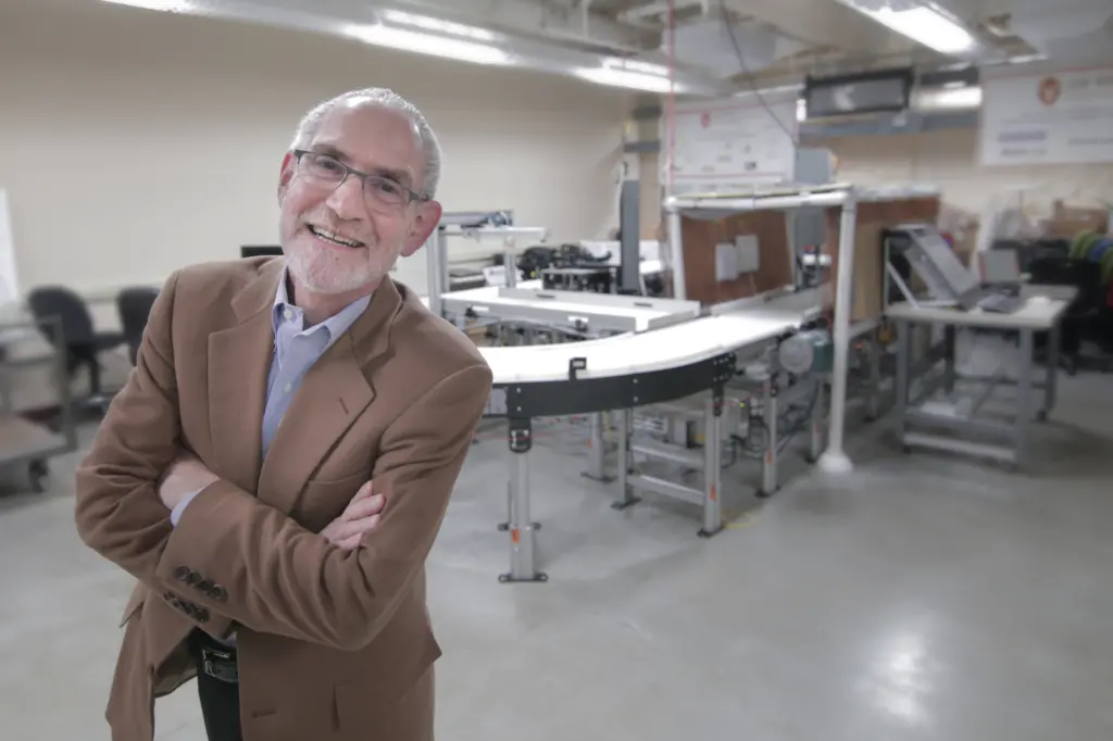 smiling man in sport jacket standing with arms crossed, standing in front of a conveyor system in an industrial laboratory