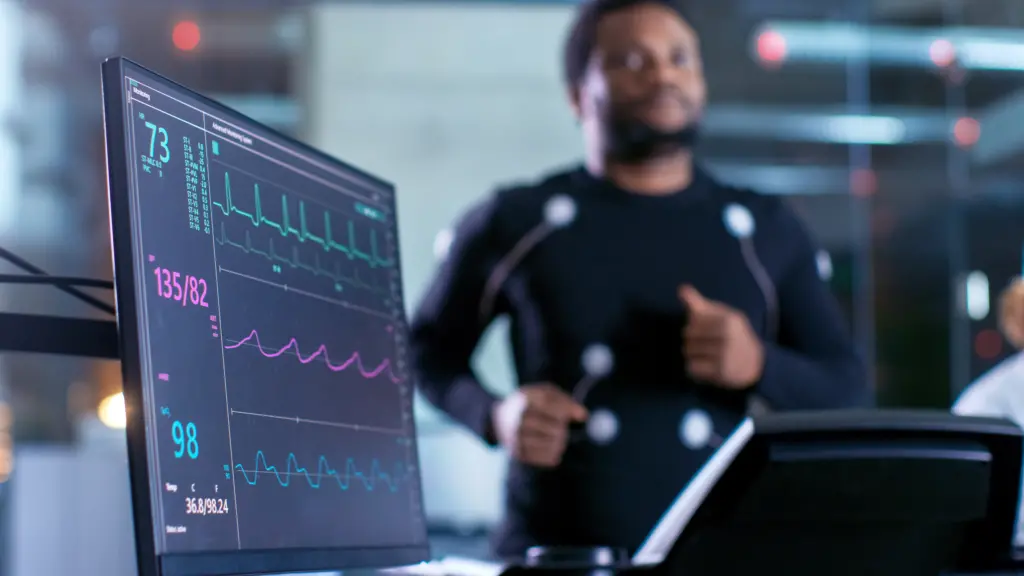 Man in background running on treadmill with computer screen in foreground showing vitals.