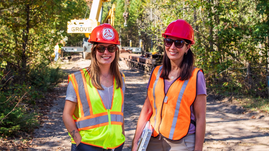 Two women wearing fluorescent safety vests and red hard hats in a wooded area with a crane behind them.