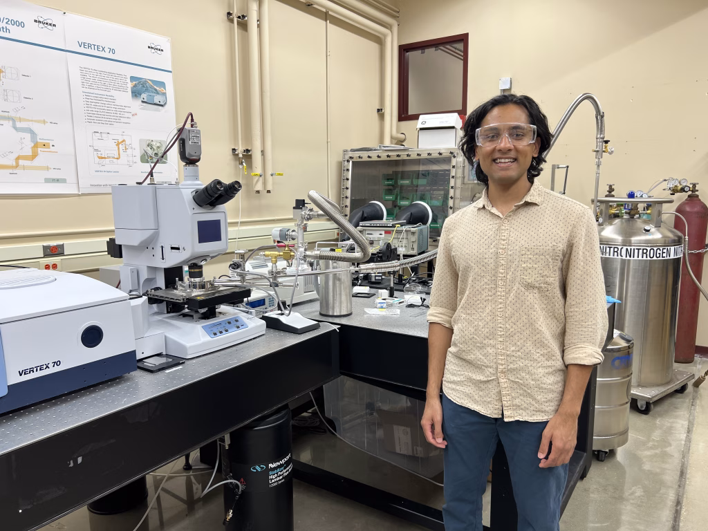 Tanuj Kumar, wearing safety glasses, a beige patterned shirt, and blue pants stands in a laboratory next to scientific equipment.