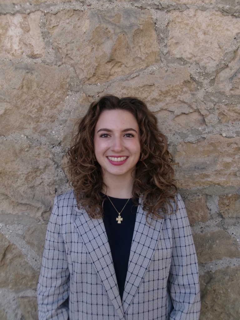 Profile of Janna Postma. She has brown curly hair and is wearing a patterned blazer.