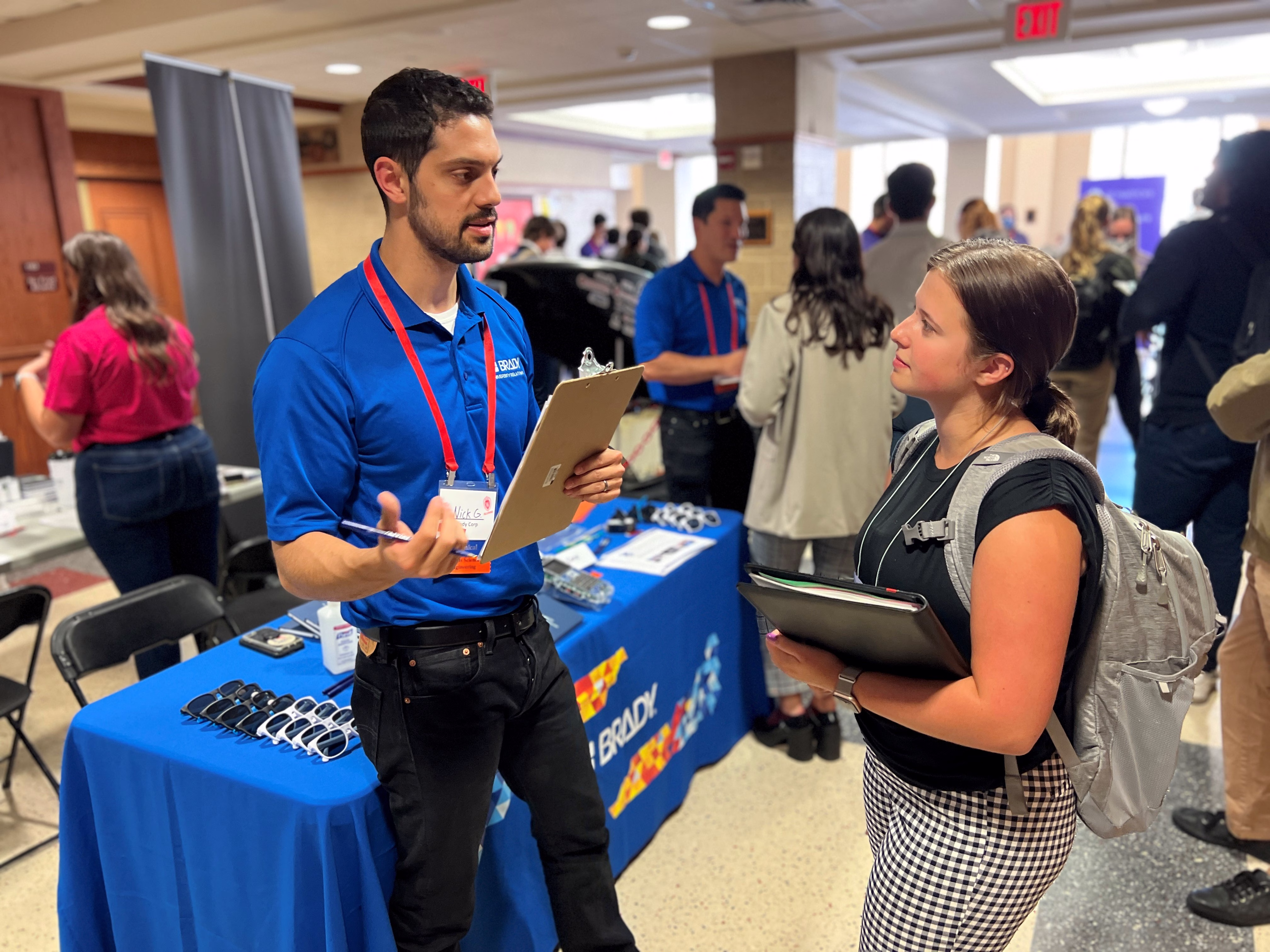 Early career man standing with a clipboard discussing opportunities with a slightly younger woman standing in front of a table with branded merchandise on it