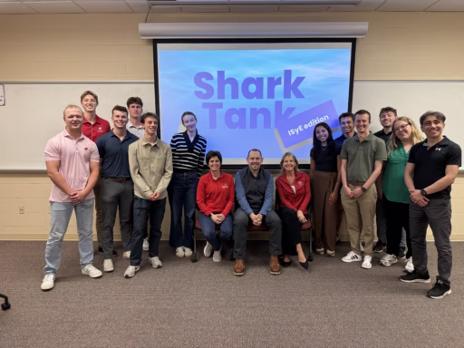 A group of college students surround three seated adult professionals with a graphic behind them
